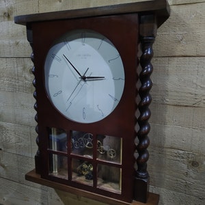 May include: A dark wood wall clock with a white face and black hands. The clock has a glass window that shows the inner workings of the clock. The clock is hanging on a wood wall.