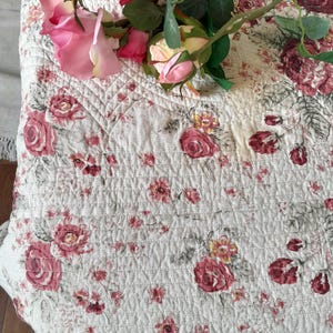 May include: A white quilted fabric with a pink floral pattern. The fabric has a subtle texture and is decorated with a bouquet of pink roses.