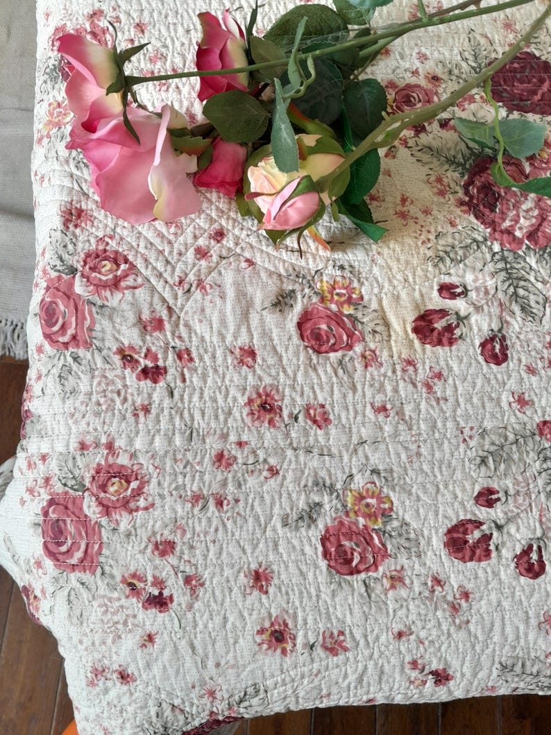 May include: A white quilted fabric with a pink floral pattern. The fabric has a subtle texture and is decorated with a bouquet of pink roses.