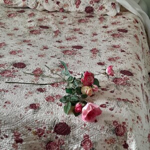 May include: A white bedspread with a floral pattern in shades of pink and burgundy. A bouquet of pink roses with green leaves is lying on the bedspread.