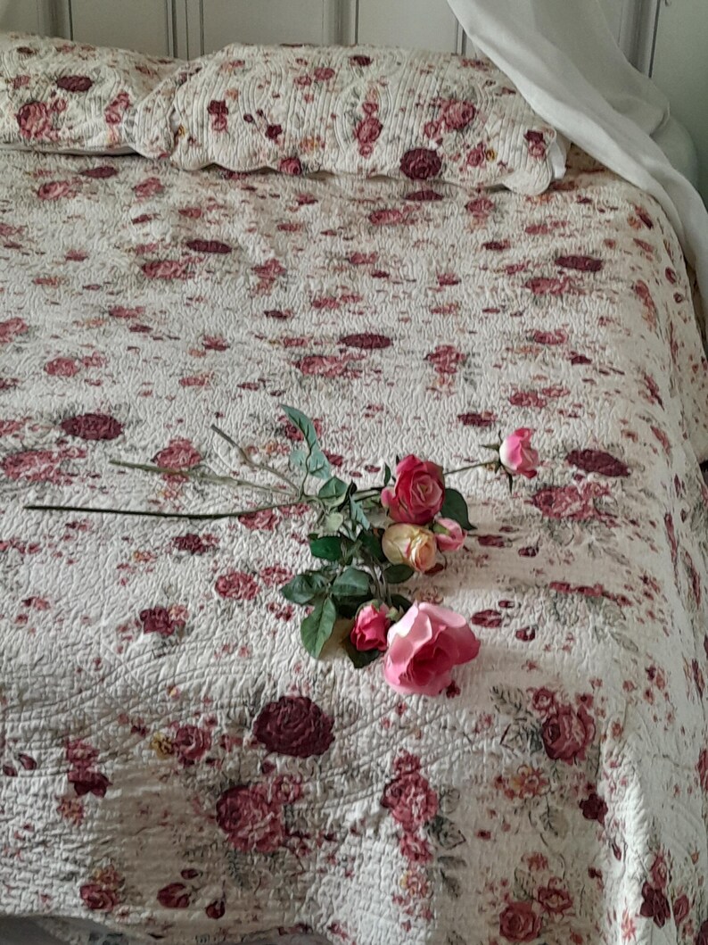 May include: A white bedspread with a floral pattern in shades of pink and burgundy. A bouquet of pink roses with green leaves is lying on the bedspread.