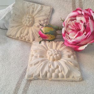May include: Two white plaster wall plaques with a raised floral design. The plaques are square and have a raised center with a flower and leaves. The plaques are on a white fabric background with a pink rose.