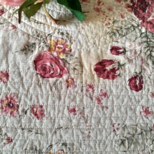 May include: A white quilted fabric with a floral pattern featuring pink and red roses. The fabric has a subtle texture and a delicate design.
