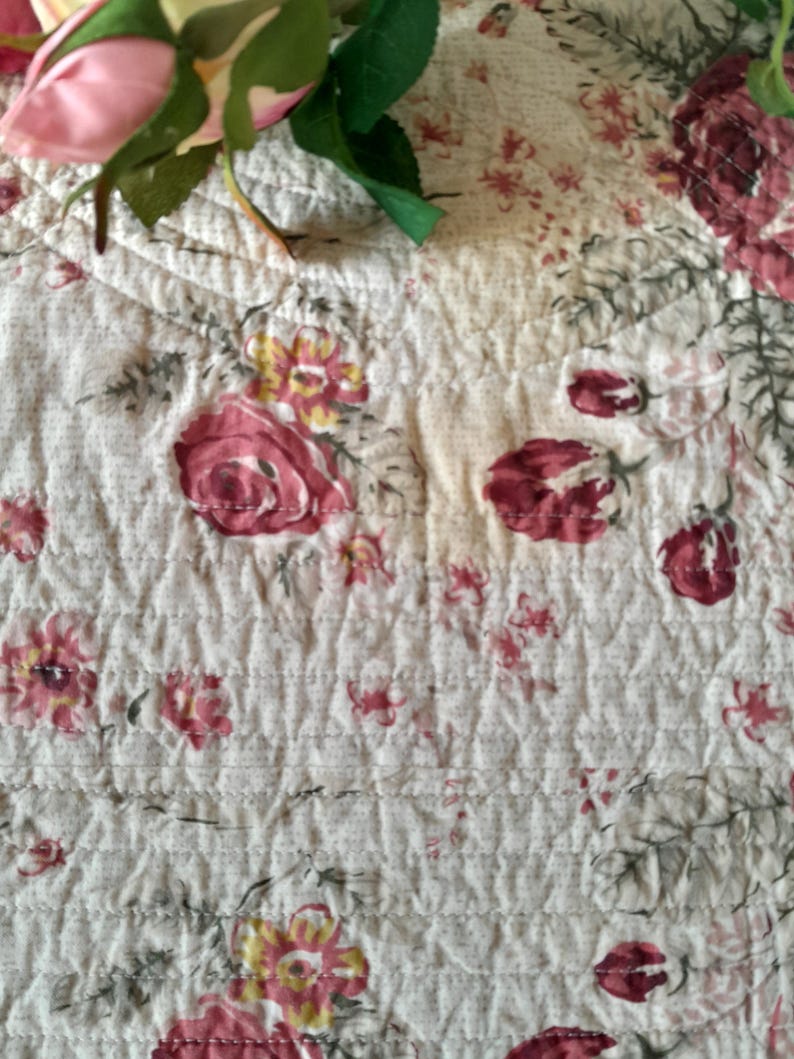 May include: A white quilted fabric with a floral pattern featuring pink and red roses. The fabric has a subtle texture and a delicate design.