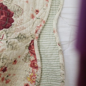 May include: A cream-colored quilted throw blanket with a floral pattern in shades of pink, red, and green. The blanket has a green and white striped border.