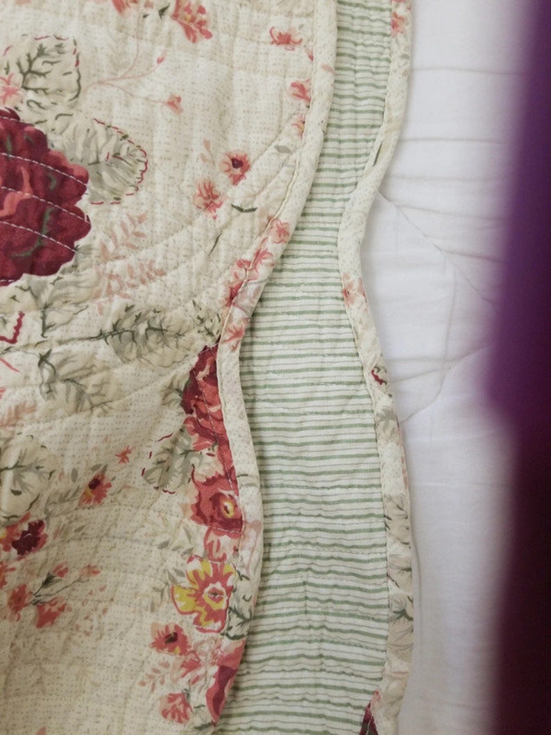 May include: A cream-colored quilted throw blanket with a floral pattern in shades of pink, red, and green. The blanket has a green and white striped border.