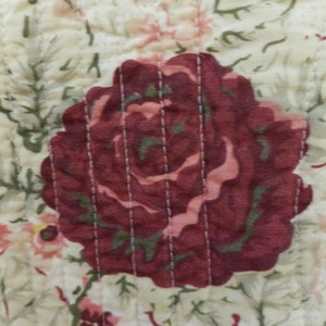 May include: A close-up of a quilted fabric with a floral pattern. The fabric features a large red rose with a pink center and green leaves. The background is a cream color with smaller pink and white flowers.
