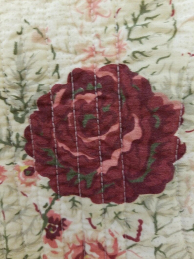 May include: A close-up of a quilted fabric with a floral pattern. The fabric features a large red rose with a pink center and green leaves. The background is a cream color with smaller pink and white flowers.