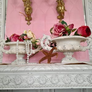 May include: Two white urns filled with pink and cream roses, adorned with a pearl necklace and a starfish. The urns sit on a white, ornate shelf against a pink backdrop, with gold cherub sconces.