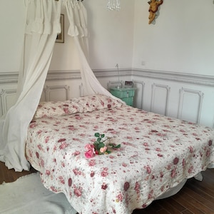 May include: A white canopy bed with a floral patterned quilt and a white rug. The bed is in a room with white walls and a chandelier.