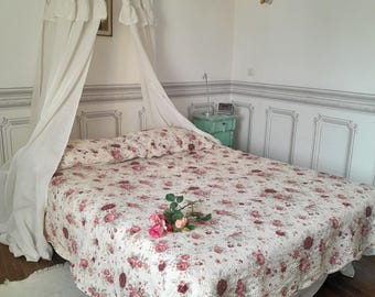 Puede incluir: Una cama con dosel blanco con un edredón estampado floral y una alfombra blanca. La cama está en una habitación con paredes blancas y una lámpara de araña.