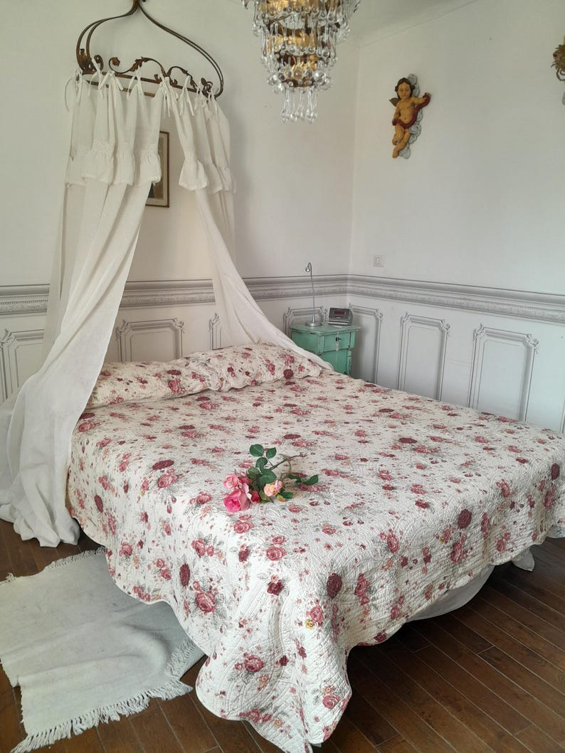 May include: A white canopy bed with a floral patterned quilt and a white rug. The bed is in a room with white walls and a chandelier.