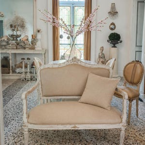 Op de afbeelding: Een beige tweezitsbank met bijpassend kussen in een kamer met een kroonluchter en een patroonvloer. De bank heeft een wit, sierlijk houten frame. Een vaas met roze bloemen is zichtbaar op de achtergrond.