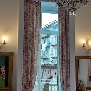 Könnte beinhalten: Innenaufnahme eines Raumes mit einem Fenster, das von rot-weiß gemusterten Vorhängen umrahmt wird. Das Fenster bietet einen Blick auf eine Stadtstraße. Goldgerahmte Spiegel und Wandleuchten mit brennenden Kerzen flankieren das Fenster. Ein Kristallleuchter hängt darüber.