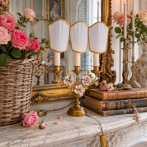 May include: A gold-toned candelabra lamp with three white pleated shades and floral accents. The lamp sits on a marble surface, next to a basket of pink roses and antique books. The lamp's cord has an on/off switch.
