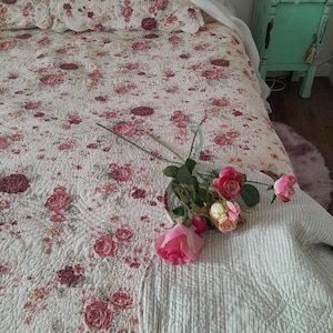May include: A white and pink floral patterned quilt with a white and gray striped throw blanket on top. A bouquet of pink roses is placed on the blanket.