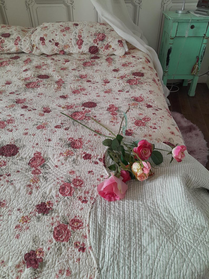 May include: A white and pink floral patterned quilt with a white and gray striped throw blanket on top. A bouquet of pink roses is placed on the blanket.