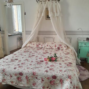 May include: A white canopy bed with a floral patterned quilt and a single pink rose on the bed. The bed is in a room with white walls and a chandelier hanging from the ceiling.