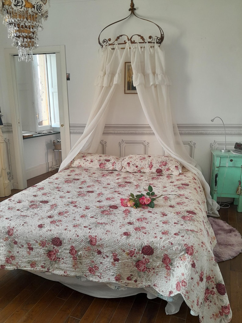 May include: A white canopy bed with a floral patterned quilt and a single pink rose on the bed. The bed is in a room with white walls and a chandelier hanging from the ceiling.