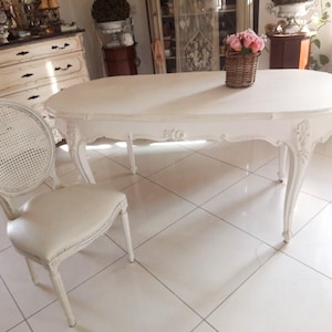 Puede incluir: Mesa ovalada blanca con un diseño tallado y una silla de mimbre blanca. La mesa está en una habitación con suelo de baldosas blancas.