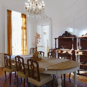 Puede incluir: Una mesa de comedor de madera con un acabado rústico y un camino de mesa de arpillera. Seis sillas rodean la mesa, con dos sillas adicionales visibles en el fondo. La habitación cuenta con un espejo grande con una corona, dos armarios antiguos y una lámpara de araña.