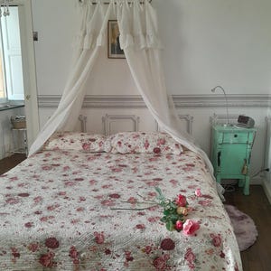 May include: A white canopy bed with a floral patterned quilt and a single rose on the bed. The bed is in a room with white walls and a green nightstand.