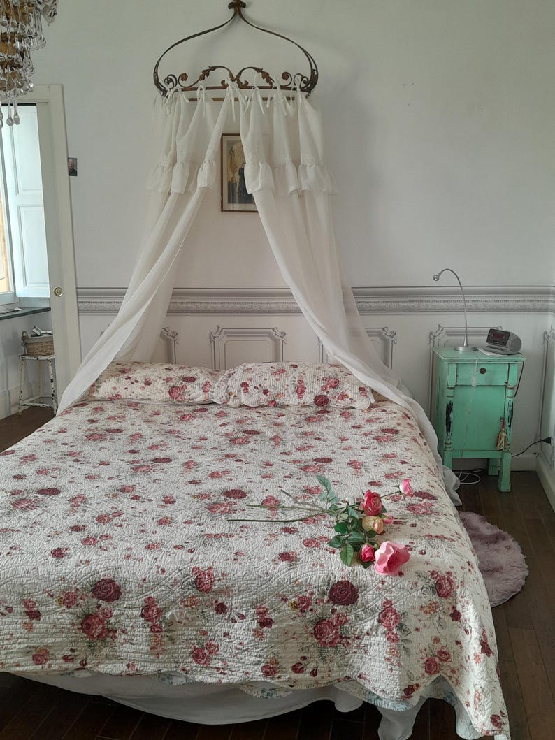 May include: A white canopy bed with a floral patterned quilt and a single rose on the bed. The bed is in a room with white walls and a green nightstand.