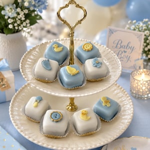 May include: A two-tiered white scalloped serving stand with a gold metal handle, holding eight square petit fours decorated with light blue and white fondant. The cakes feature small yellow and blue edible toppers shaped like ducks, buttons, and feet.