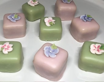 May include: Nine square-shaped petit fours with pastel green and pink icing. Each petit four is decorated with a single sugar flower in various colors, including pink, blue, and white.