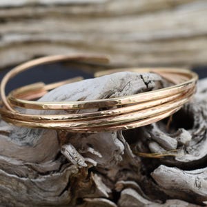 May include: A set of three interlocking bangles crafted from polished rose gold metal. Each bangle features a distinct hammered texture across its surface, creating a reflective finish. The circular bands are connected in a continuous loop design.