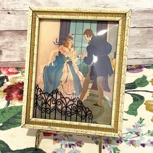 May include: A framed vintage print depicting a couple in period attire. The print features a woman in a blue gown and a man in a dark coat. The frame is cream-colored with gold accents, and the print is displayed on a gold-toned stand.