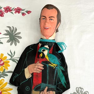 May include: A vintage fabric doll depicting a man in a black suit with a red plaid vest, holding a top hat with a colorful parrot. The doll has a painted face and is set against a floral patterned background.