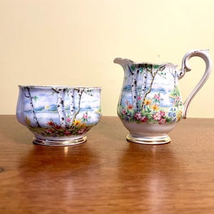 May include: A porcelain creamer and sugar bowl set featuring a painted landscape scene with white birch trees, colorful wildflowers, and a blue lake. Both pieces have a scalloped rim and a gold-trimmed base, with the creamer having a curved handle.