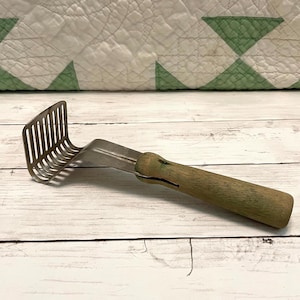 May include: A vintage potato masher with a stainless steel head and a wooden handle. The masher features a rectangular head with vertical tines and a curved design. The wooden handle is a light brown color with green accents.