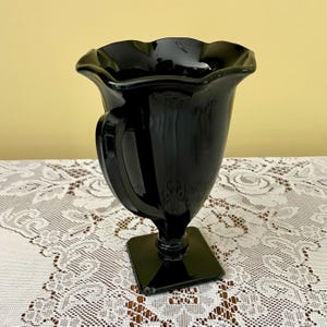 Peut inclure: Un vase en verre noir brillant dot&eacute; d'un large bord festonn&eacute; et ondul&eacute; ainsi que d'un corps lisse et incurv&eacute;. Le vase est soutenu par une tige cylindrique courte qui se connecte &agrave; une base carr&eacute;e, offrant une assise stable au r&eacute;cipient.