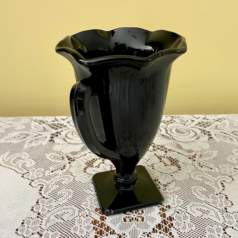 Peut inclure: Un vase en verre noir brillant dot&eacute; d'un large bord festonn&eacute; et ondul&eacute; ainsi que d'un corps lisse et incurv&eacute;. Le vase est soutenu par une tige cylindrique courte qui se connecte &agrave; une base carr&eacute;e, offrant une assise stable au r&eacute;cipient.
