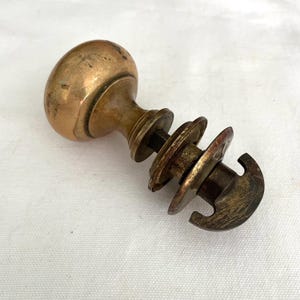 May include: Antique brass door knob with a rounded handle and a decorative stem. The knob has a weathered appearance, showing signs of age and use. The stem features a series of circular elements and a hook-shaped end.