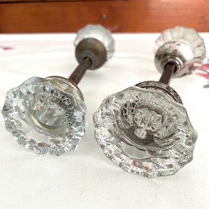 May include: Two antique glass door knobs with a clear, faceted design. The knobs have a flower-like shape and are attached to aged metal stems. The knobs are likely for a vintage or antique door.