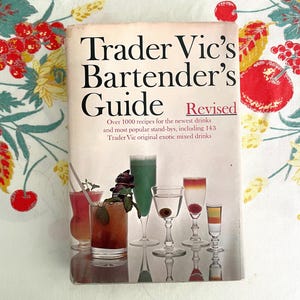 May include: A vintage copy of "Trader Vic's Bartender's Guide, Revised" with a white cover and black text. The cover features images of colorful cocktails in various glasses. The book promises over 1000 drink recipes.