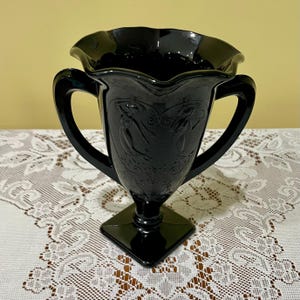 Peut inclure: Un vase en verre noir avec un bord ondul&eacute; et deux anses lat&eacute;rales. Le corps du vase pr&eacute;sente un motif en relief moul&eacute; repr&eacute;sentant deux silhouettes dans un cadre en forme de c&oelig;ur, reposant sur une base carr&eacute;e. Le verre a une finition brillante.