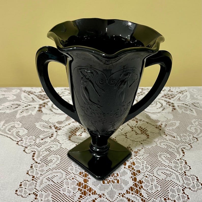 Peut inclure: Un vase en verre noir avec un bord ondul&eacute; et deux anses lat&eacute;rales. Le corps du vase pr&eacute;sente un motif en relief moul&eacute; repr&eacute;sentant deux silhouettes dans un cadre en forme de c&oelig;ur, reposant sur une base carr&eacute;e. Le verre a une finition brillante.