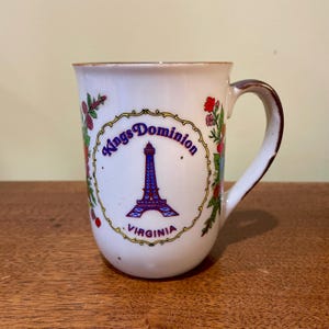 May include: A white ceramic mug featuring a blue graphic of the Eiffel Tower, the text Kings Dominion and VIRGINIA, and a decorative yellow border. The mug is decorated with colorful floral and berry illustrations on both sides of the central design.