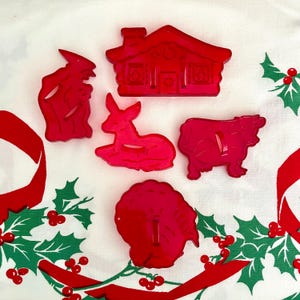 May include: A set of five red plastic cookie cutters featuring a house, a standing figure, a deer, a cow, and a turkey. The cutters are arranged on a white fabric surface decorated with a green and red holly leaf and berry pattern.