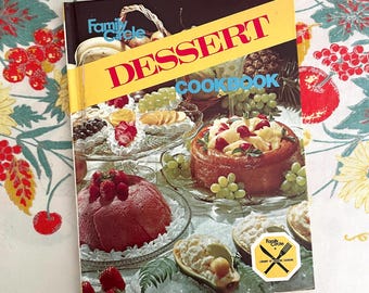 May include: A vintage "Family Circle Dessert Cookbook" with a yellow banner reading "DESSERT COOKBOOK." The cover features images of various desserts, including cakes and fruit arrangements. The cookbook is on a floral tablecloth.