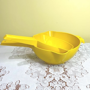 May include: A bright yellow plastic kitchen bowl with a matching yellow plastic scoop nested inside. Both items feature long, sturdy handles with a small hole at the end for hanging. The bowl has a wide, rounded basin and a smooth, solid finish.