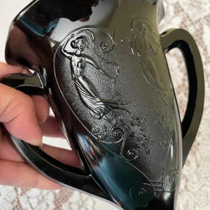 Peut inclure: Un pichet en verre noir pr&eacute;sentant un relief repr&eacute;sentant deux personnages v&ecirc;tus de v&ecirc;tements fluides entour&eacute;s de motifs d&eacute;coratifs tourbillonnants. Le r&eacute;cipient poss&egrave;de un large bec verseur et une anse incurv&eacute;e, avec une finition lisse et brillante.
