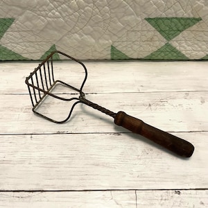 May include: An antique potato masher with a dark wooden handle and a twisted metal shaft. The masher head is made of a grid of metal bars. The kitchen utensil is resting on a white wooden surface.