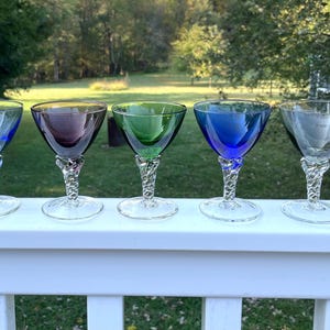 May include: Five colorful cocktail glasses with twisted stems. The glasses are in shades of blue, purple, green, and clear. They are arranged in a row on a white surface, with a blurred green background of trees and grass.
