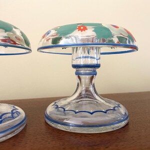May include: Two clear glass candle holders with a mushroom-shaped top. The tops are decorated with a floral pattern in teal, red, and white. The base and rim are outlined in blue. The candle holders are on a brown surface.