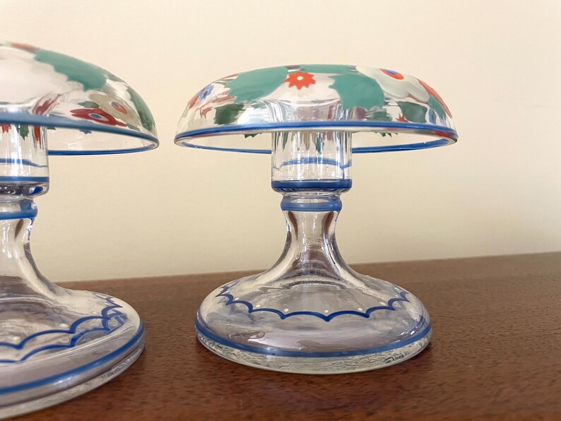 May include: Two clear glass candle holders with a mushroom-shaped top. The tops are decorated with a floral pattern in teal, red, and white. The base and rim are outlined in blue. The candle holders are on a brown surface.